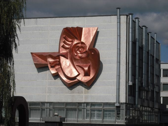 Vemos una edificación donde hay una escultura pegada a la pared, una paloma en su pico tiene un átomo