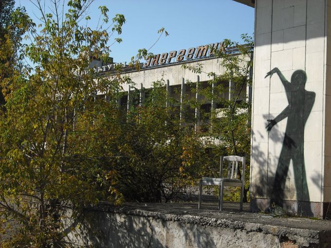 Tenemos  una pared donde vemos un graffiti de una figura en negro, también sillas un poco destruidas y también abundante vegetación