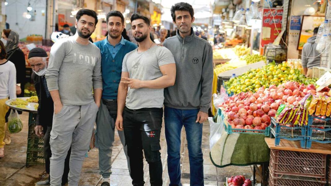 Cuatro hombres jóvenes posan juntos en un mercado concurrido, junto a puestos con granadas, limones y otras frutas, con compradores alrededor.