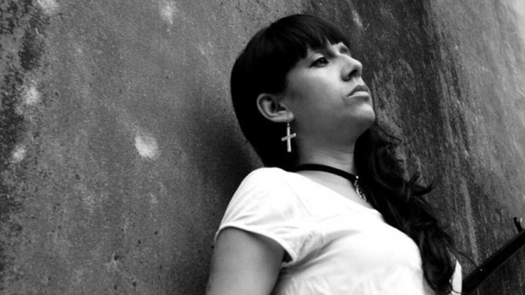 Mujer joven de cabello oscuro con flequillo, camiseta clara y arete de cruz, apoyada contra una pared texturizada en blanco y negro.