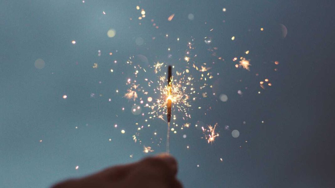 Una mano sostiene una bengala encendida que despide chispas doradas y puntos de luz desenfocados sobre un fondo azul grisáceo.