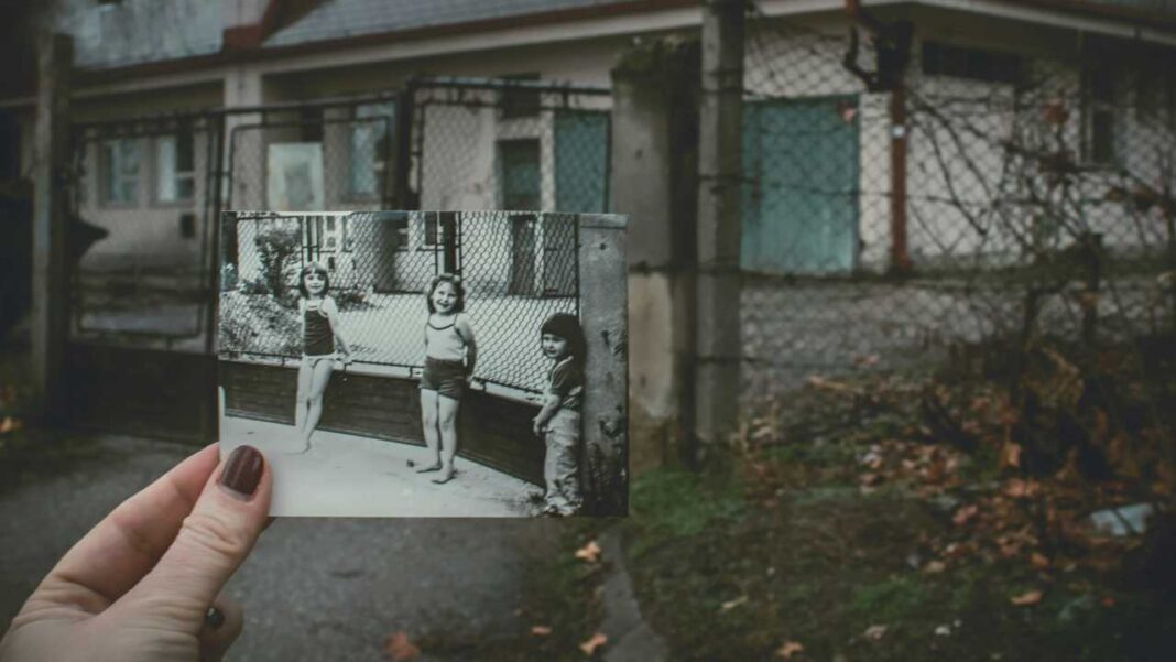 Una mano con uñas pintadas sostiene una fotografía en blanco y negro de tres niñas junto a una valla, delante de un edificio y un patio con hojas secas.