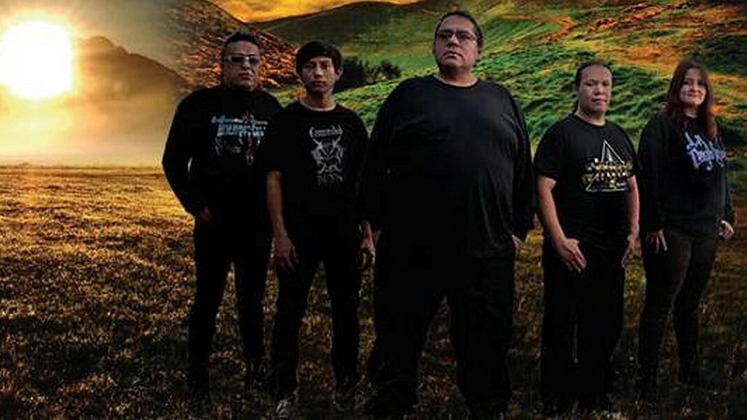 Cinco personas vestidas de negro posan de pie en un campo abierto con hierba, colinas al fondo y un sol brillante bajo en el horizonte.