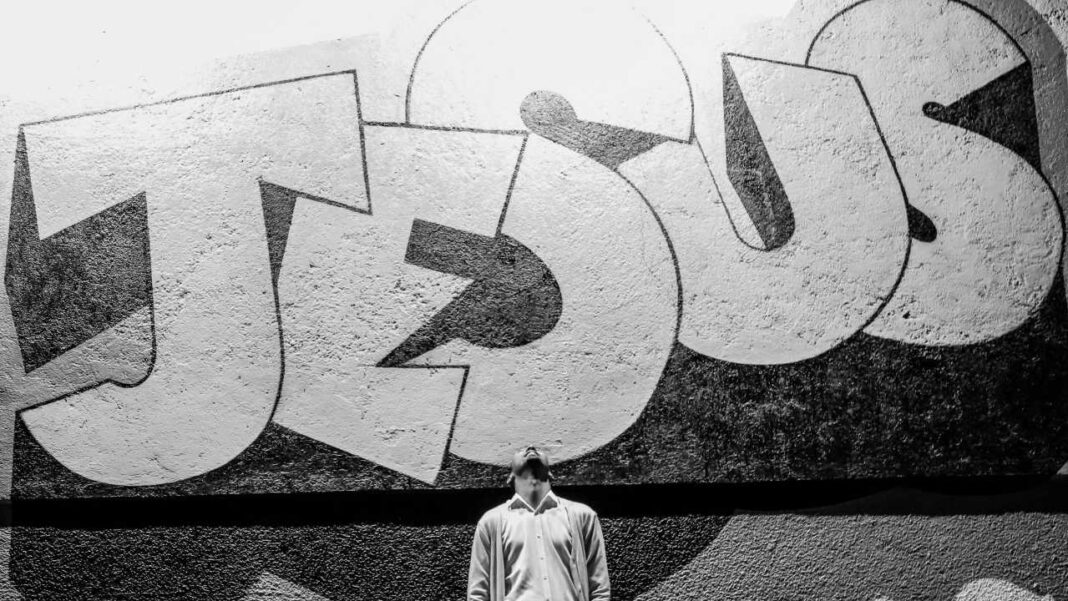 Imagen en blanco y negro de un gran grafiti con letras blancas sobre una pared texturizada; una persona con camisa clara está de pie en la parte inferior central.