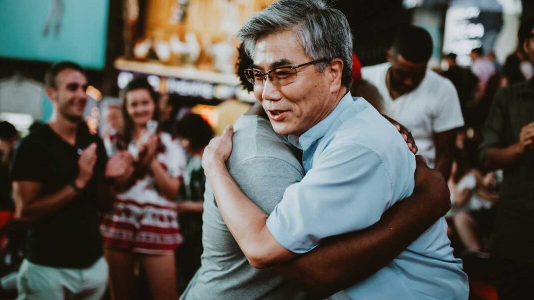 Hombre mayor de cabello gris y gafas, con camisa azul claro, abraza a otra persona mientras varias personas desenfocadas observan y aplauden al fondo.