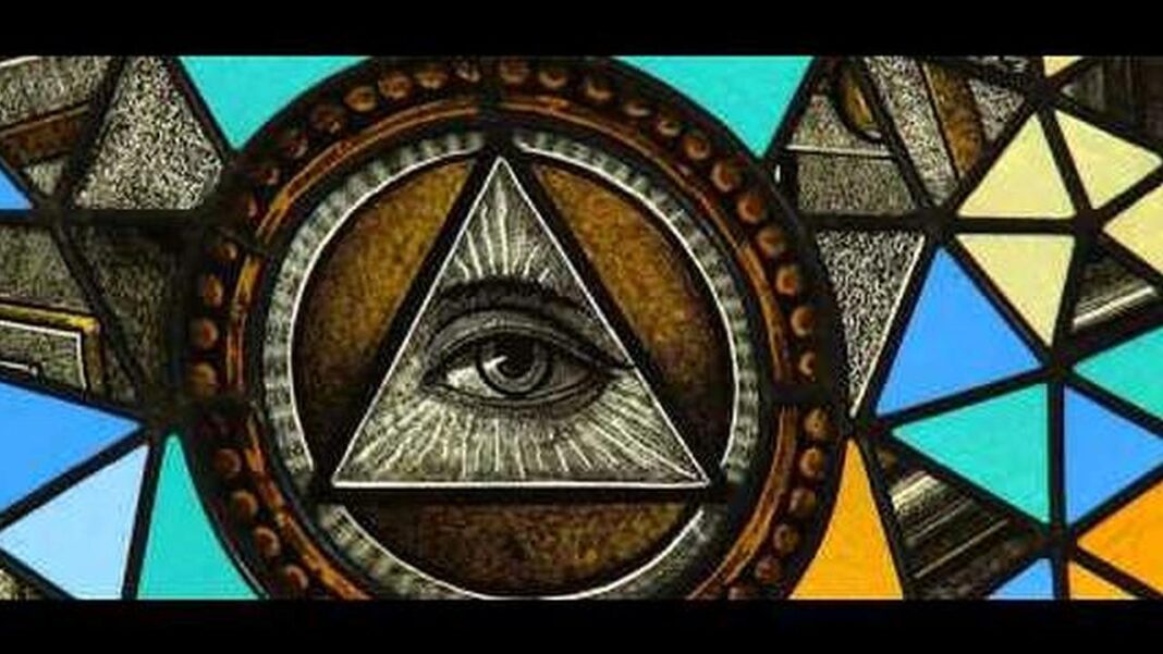 Vitral con un ojo dentro de un triángulo radiante, rodeado por un círculo decorativo y paneles geométricos de colores azules, verdes y amarillos.