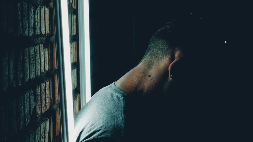 Persona de perfil en sombra junto a una ventana luminosa, con camiseta gris y mano en la cabeza, frente a una pared texturizada.