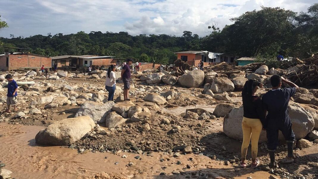 Personas de pie entre grandes rocas, lodo y restos arrastrados frente a casas de ladrillo, con colinas boscosas y cielo nublado al fondo.