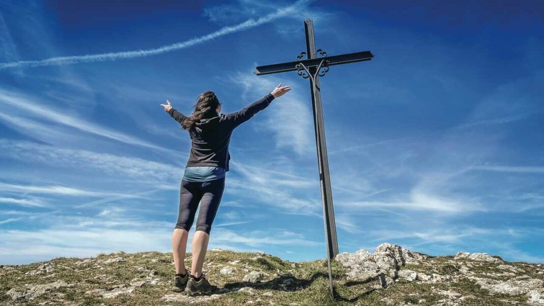 Una persona de espaldas con los brazos abiertos frente a una gran cruz de metal, sobre un terreno rocoso bajo un cielo azul con nubes finas.