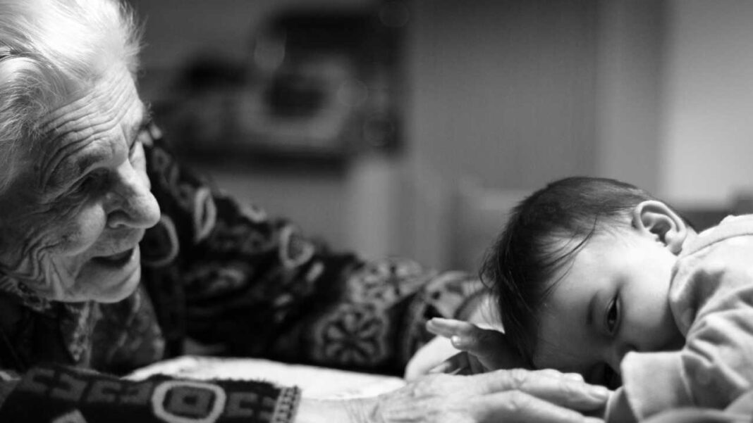 Fotografía en blanco y negro de una anciana y un bebé mirándose de cerca; ella sonríe y él está recostado, con el fondo desenfocado.