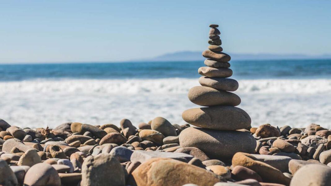 Una torre de piedras lisas apiladas en equilibrio sobre una playa llena de guijarros, con olas del mar y cielo azul al fondo.