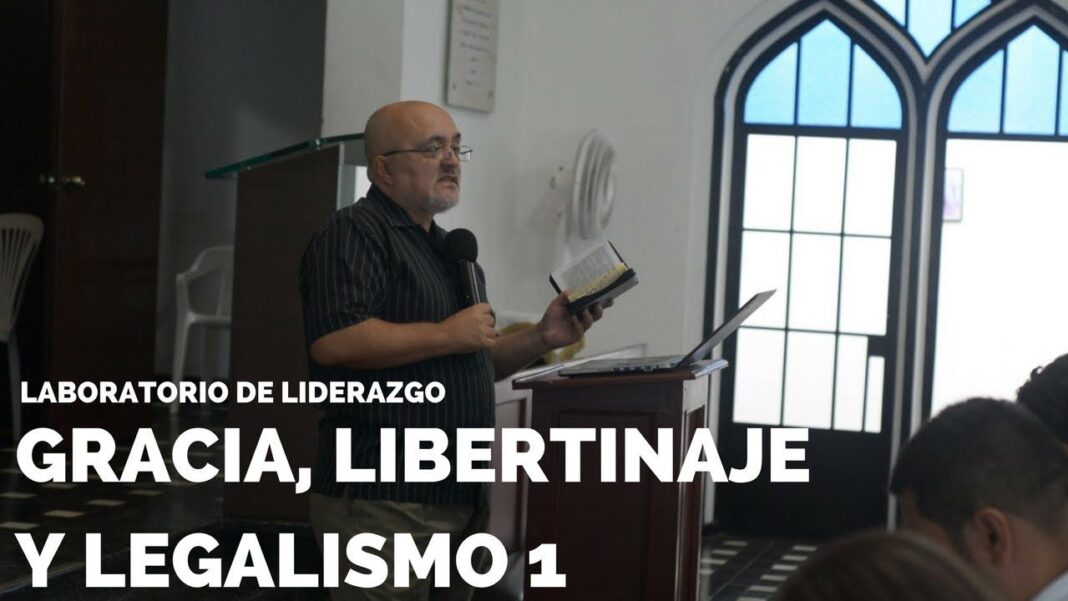 Hombre calvo con lentes y camisa a rayas sostiene un micrófono y un libro junto a un atril con computadora, frente a ventanas arqueadas y público.