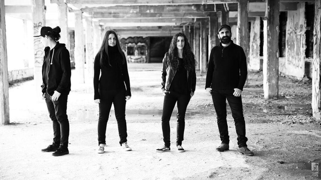Cuatro personas vestidas de negro están de pie en un edificio abandonado con columnas y paredes deterioradas, en una fotografía en blanco y negro.
