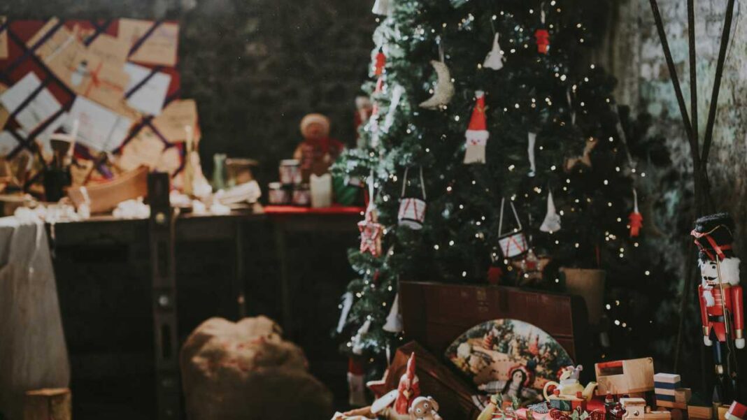 Árbol de Navidad iluminado con adornos junto a un cascanueces de madera, una caja abierta con objetos decorativos y un saco en una habitación rústica.