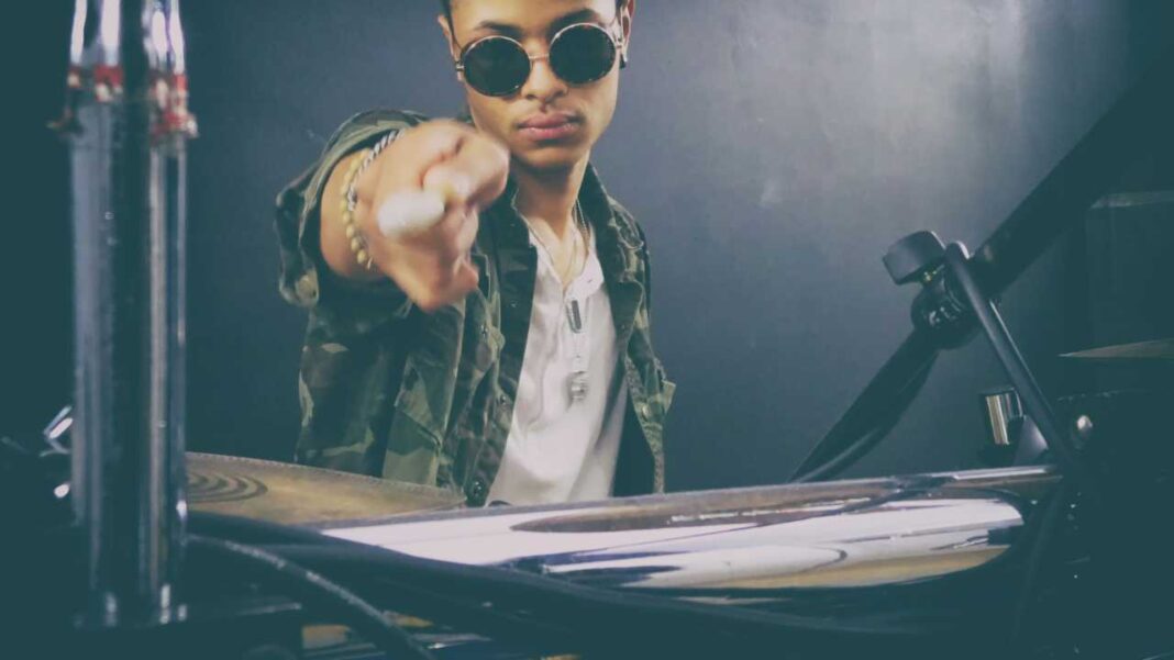 Una persona con gafas redondas y chaqueta de camuflaje señala hacia la cámara junto a equipo de percusión sobre fondo oscuro.