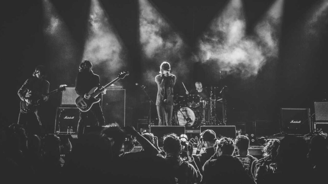 Concierto en blanco y negro: cantante con micrófono al centro, guitarristas a la izquierda, batería al fondo, luces y humo sobre el escenario, público en silueta.