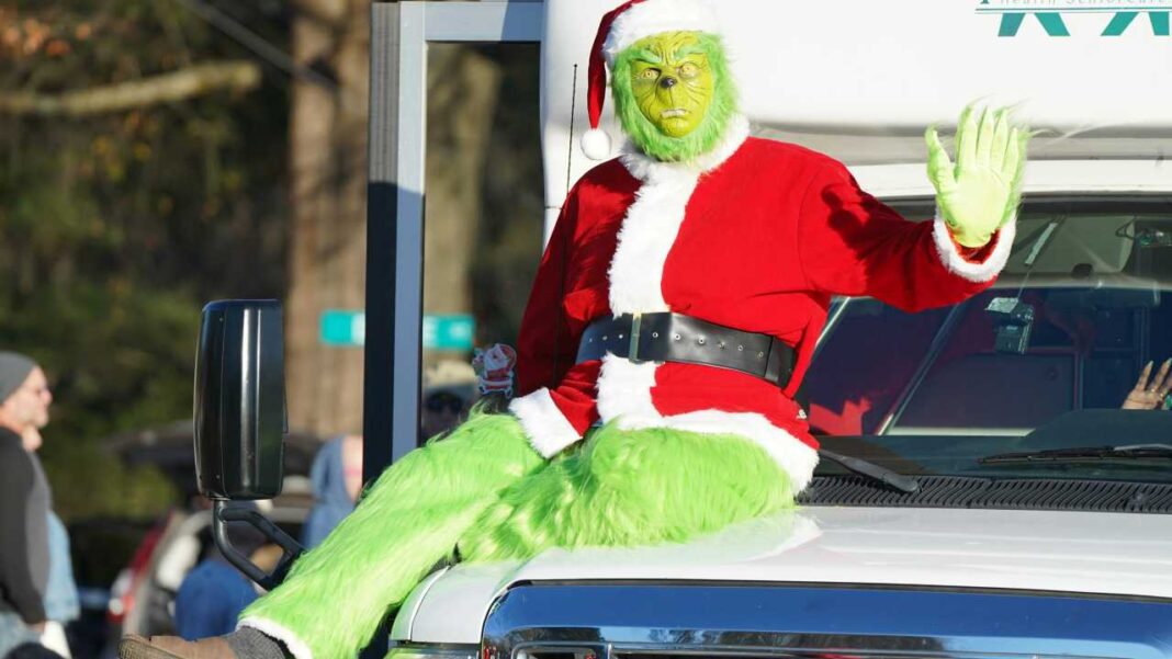 Persona con máscara verde y traje rojo de Papá Noel sentada en el capó de un vehículo blanco, saludando con la mano derecha.