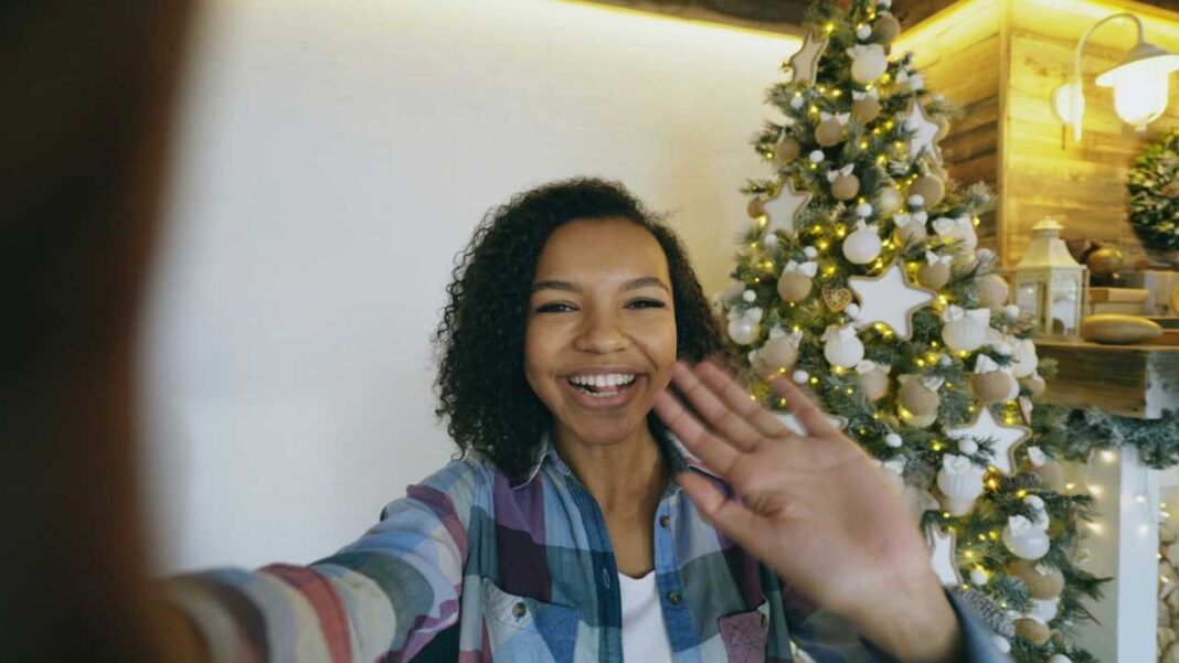 Mujer joven de cabello rizado y camisa a cuadros sonríe y saluda con la mano frente a un árbol de Navidad iluminado junto a una pared blanca.