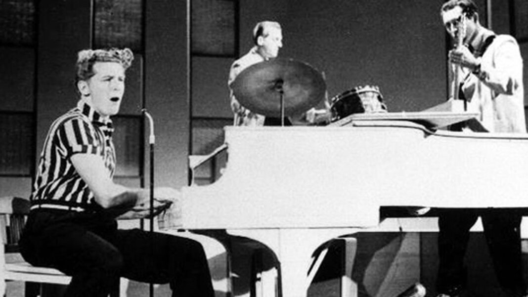 En blanco y negro, tres músicos en un escenario: un hombre canta y toca el piano, otro la batería y un tercero está de pie con un micrófono.