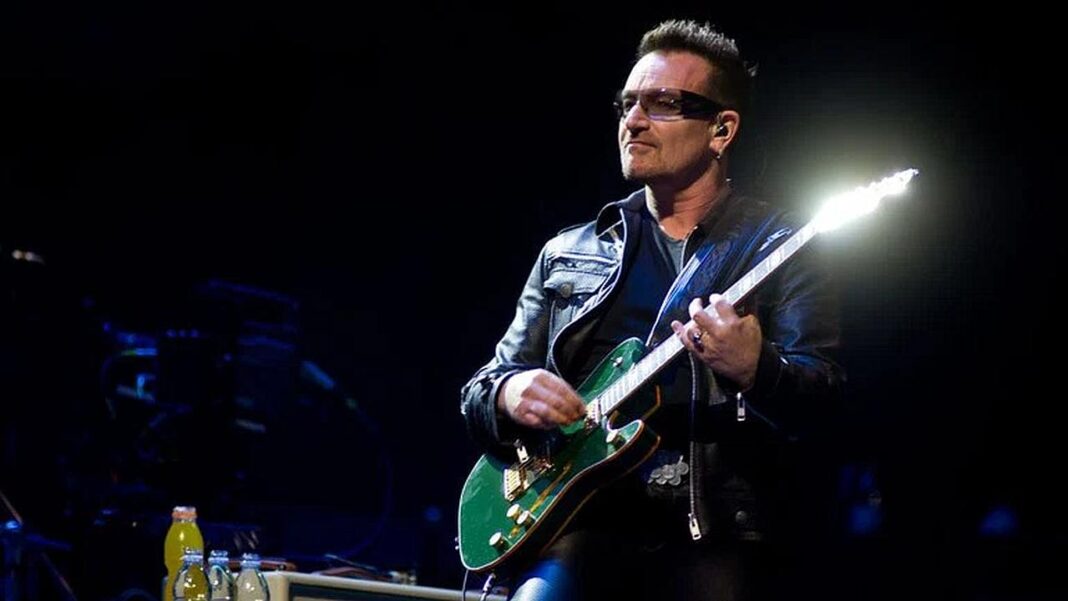 Hombre con gafas oscuras y chaqueta de cuero negra toca una guitarra eléctrica verde en un escenario oscuro iluminado.