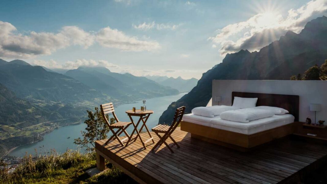 Cama al aire libre sobre una plataforma de madera con dos sillas y una mesa pequeña, frente a un lago, montañas y un cielo con sol radiante.