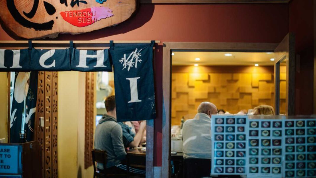 Entrada de un local de sushi con letrero de madera, cortinas azules con letras y clientes sentados en el interior iluminado.