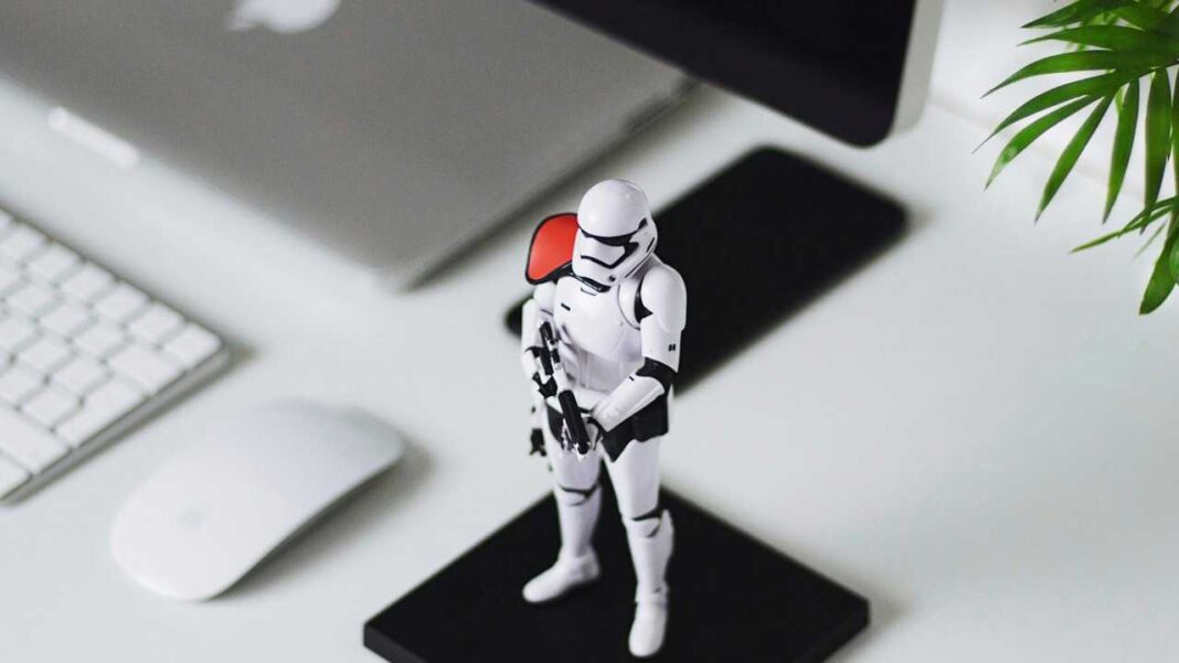 Figura de soldado blanco con armadura y hombrera roja sobre base negra, rodeada de teclado, ratón, portátil y hojas verdes en un escritorio claro.
