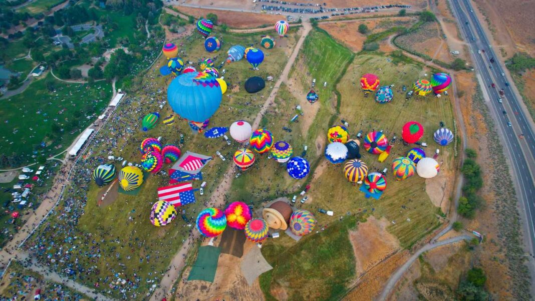 Vista aérea de numerosos globos aerostáticos de colores sobre un campo verde, con multitud de personas, vehículos y una carretera cercana.