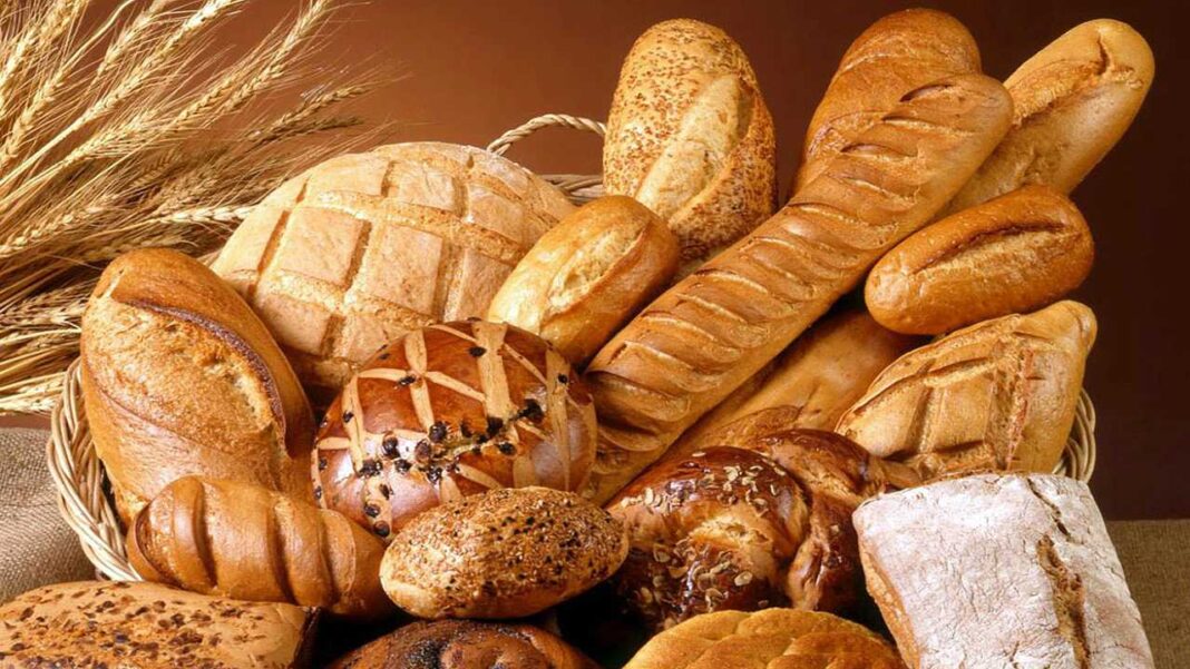 Cesta de mimbre con panes variados, hogazas, baguettes y bollos con semillas, junto a espigas de trigo sobre fondo marrón.