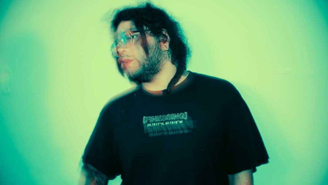 Persona joven de pelo oscuro, barba y gafas mira hacia un lado; viste camiseta negra con texto y coordenadas, sobre fondo verde turquesa iluminado.