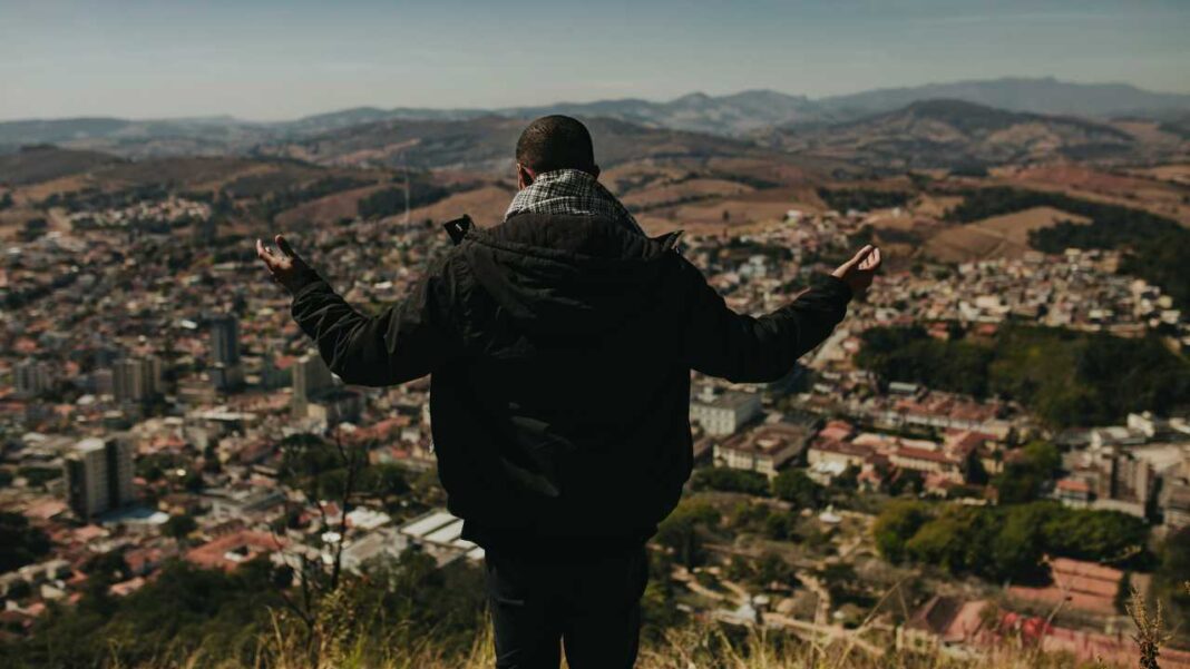 Persona de espaldas con chaqueta oscura y bufanda, brazos extendidos, contemplando una ciudad extensa rodeada de colinas y montañas bajo un cielo claro.