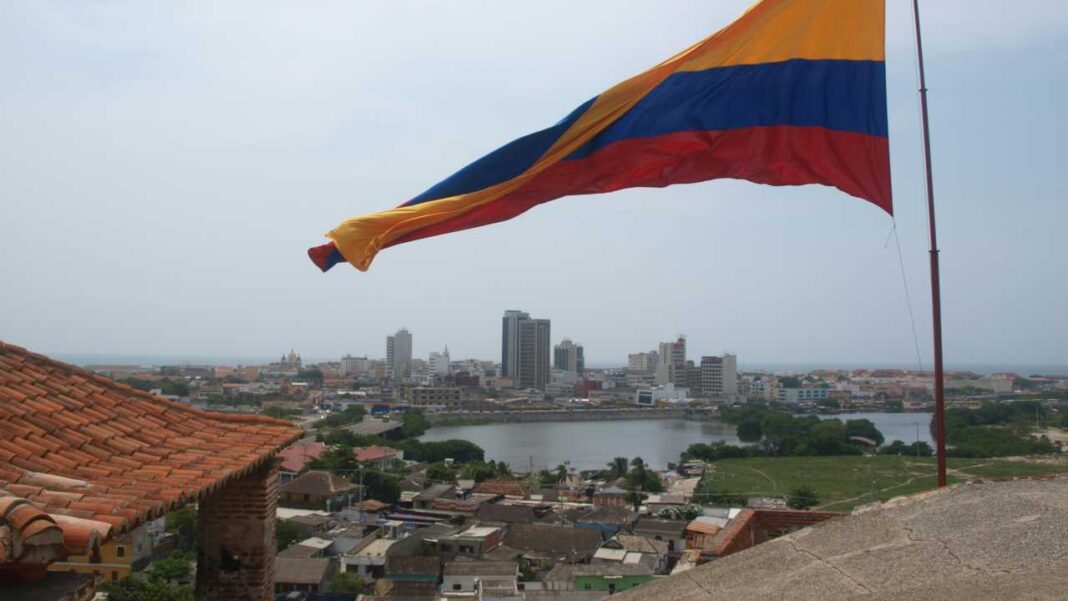 Una bandera grande de franjas amarilla, azul y roja ondea sobre una ciudad con edificios, un cuerpo de agua y tejados rojos en primer plano.