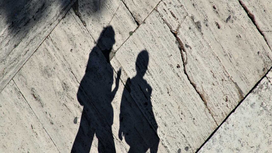 Sombras alargadas de dos personas proyectadas sobre un pavimento de piedra clara con grietas y líneas diagonales bajo luz solar.