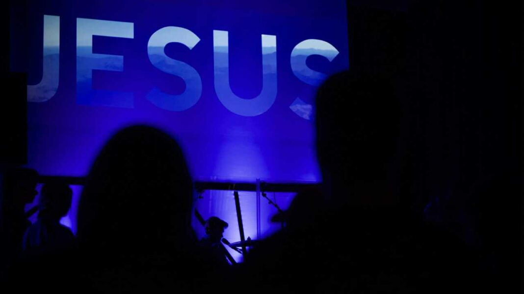 Letras grandes de la palabra JESUS iluminadas en azul sobre fondo oscuro, con siluetas de personas en primer plano.