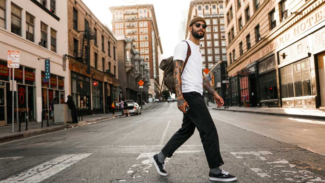 Hombre con gorro, gafas de sol, camiseta blanca, pantalón negro y mochila camina sobre un paso de cebra en una calle urbana con edificios y un coche al fondo.