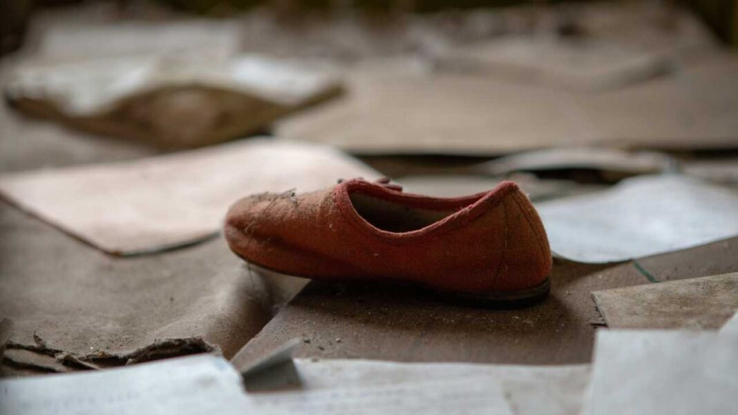 Un zapato rojo pequeño y desgastado descansa sobre cartones y papeles dispersos en el suelo, con el fondo desenfocado.