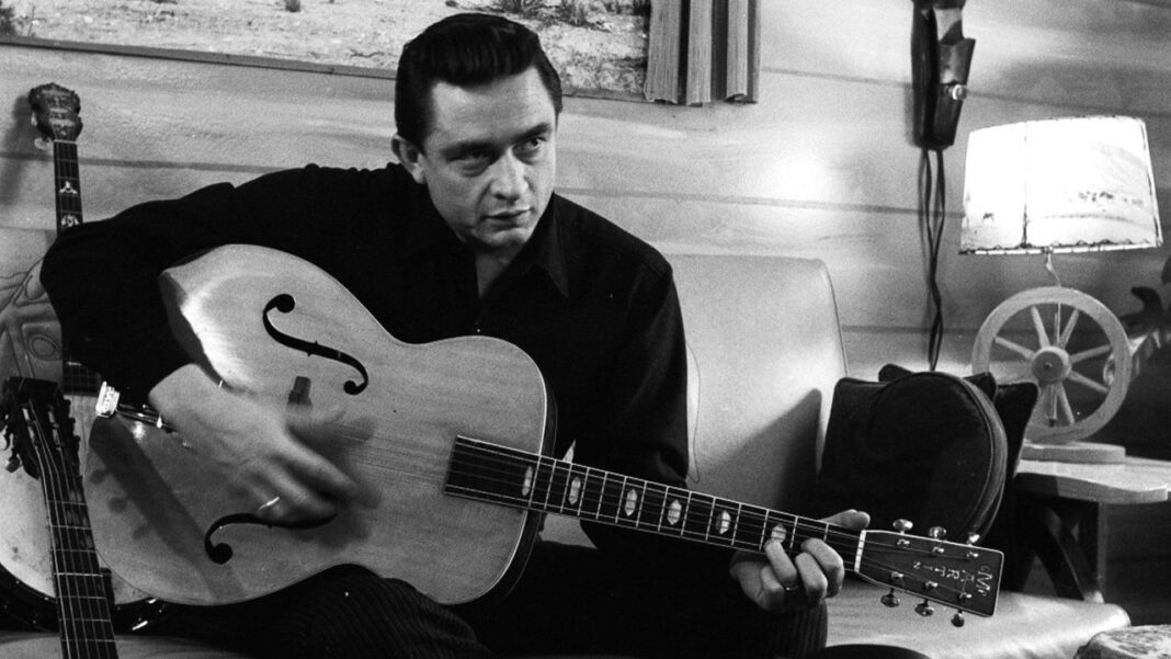 Hombre de pelo oscuro y camisa negra toca una guitarra acústica de cuerpo grande en una sala con pared de madera y lámpara.