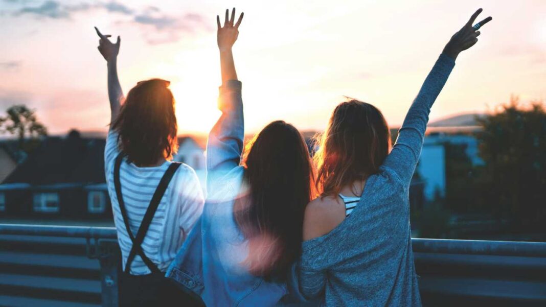 Tres jóvenes de espaldas con los brazos levantados frente a un cielo al atardecer, sobre una barandilla, con edificios y árboles al fondo.