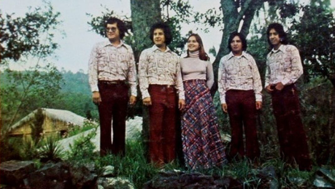 Cinco personas posan de pie al aire libre entre árboles y vegetación; cuatro hombres con camisas estampadas y pantalones oscuros, y una mujer con suéter y falda estampada.