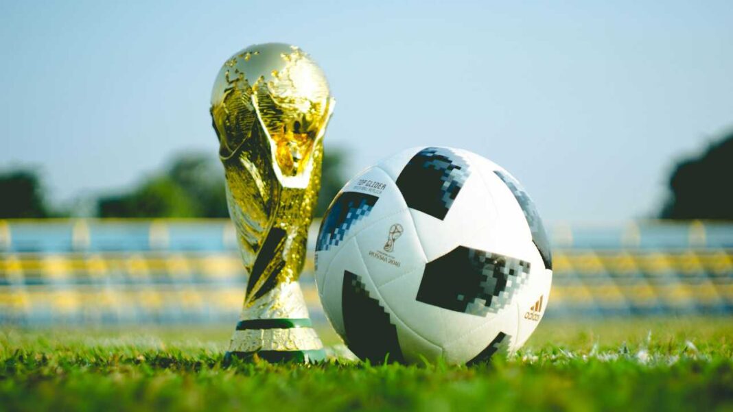 Trofeo dorado junto a un balón de fútbol blanco y negro sobre césped verde, con fondo de estadio desenfocado y cielo claro.