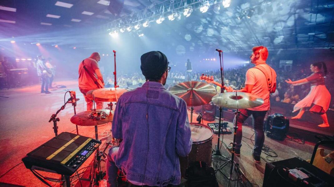 Baterista de espaldas con chaqueta vaquera y gorro negro toca la batería en un escenario iluminado, junto a guitarristas y público.