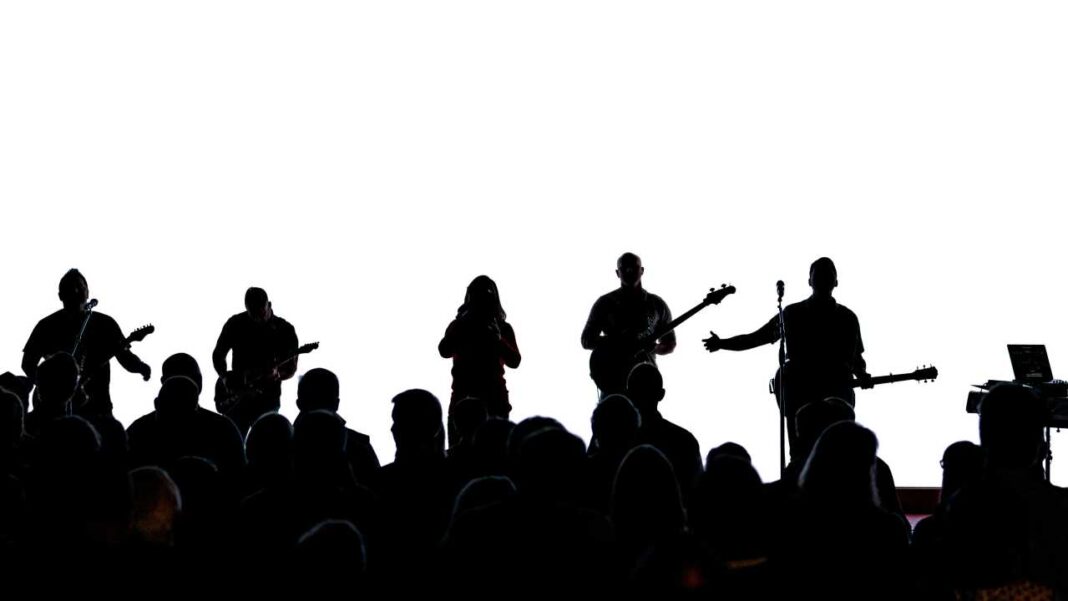 Siluetas de varios músicos con guitarras y micrófono sobre un escenario, frente a una multitud de público, sobre un fondo blanco iluminado.