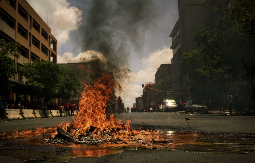 Una gran hoguera arde sobre escombros en medio de una calle urbana, con humo negro elevándose y personas y vehículos al fondo.