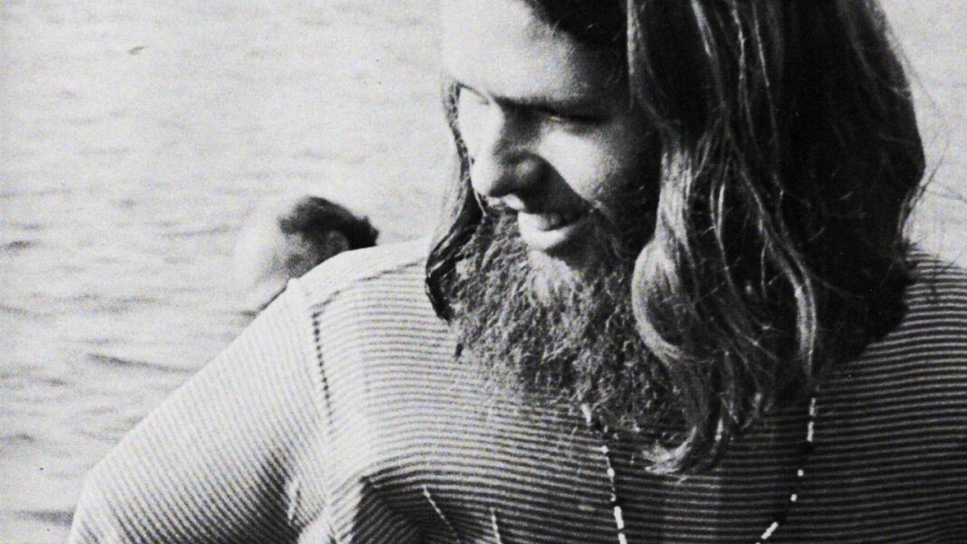 Retrato en blanco y negro de un hombre joven con barba y pelo largo, camiseta rayada y collar, mirando hacia abajo junto al agua.