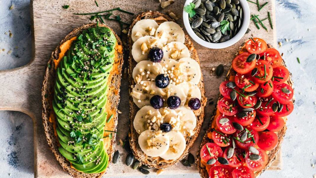 Tres tostadas sobre una tabla de madera: una con aguacate, otra con plátano y arándanos, y otra con tomates cherry y semillas.