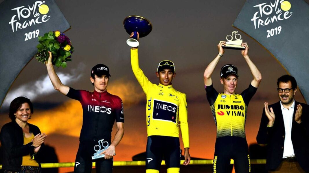 Tres ciclistas en un podio sostienen un trofeo, flores y una placa ante los logotipos del Tour de France 2019, con dos personas aplaudiendo.