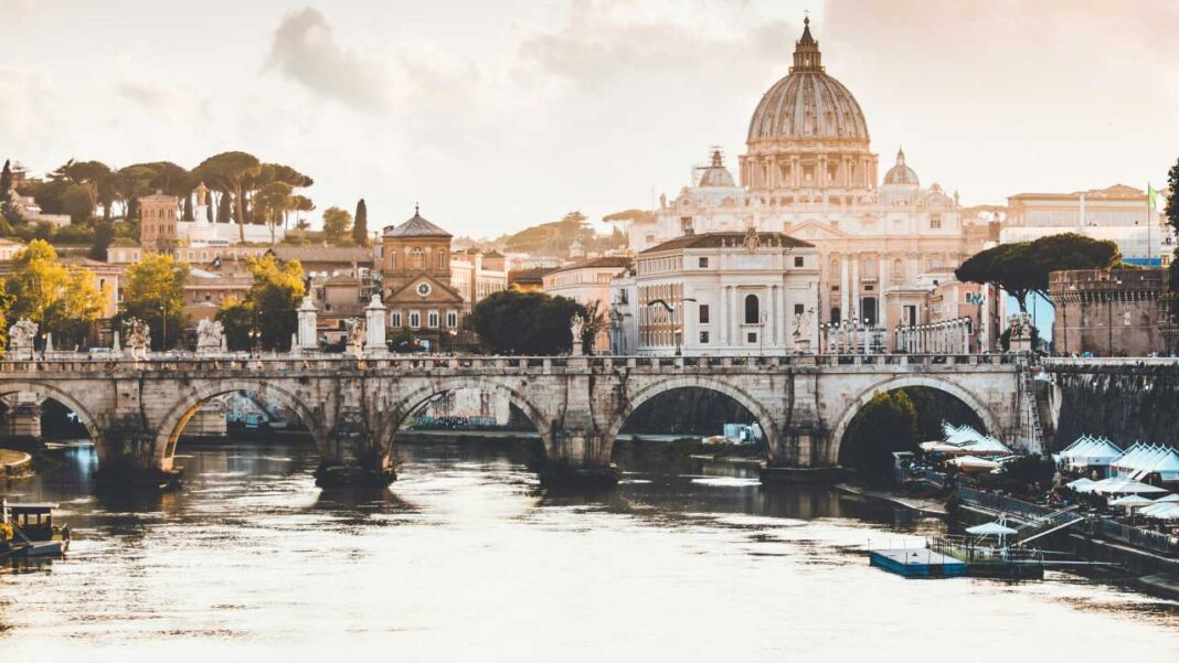 Vista de un río ancho con un puente de piedra de varios arcos y estatuas, grandes edificios con cúpula, árboles y sombrillas junto a la orilla.