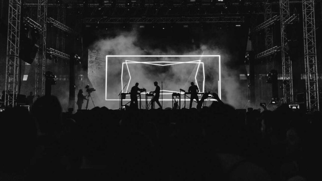 Escenario oscuro con tres músicos junto a mesas de equipo, rodeados de humo y una gran estructura luminosa rectangular, ante una multitud en silueta.