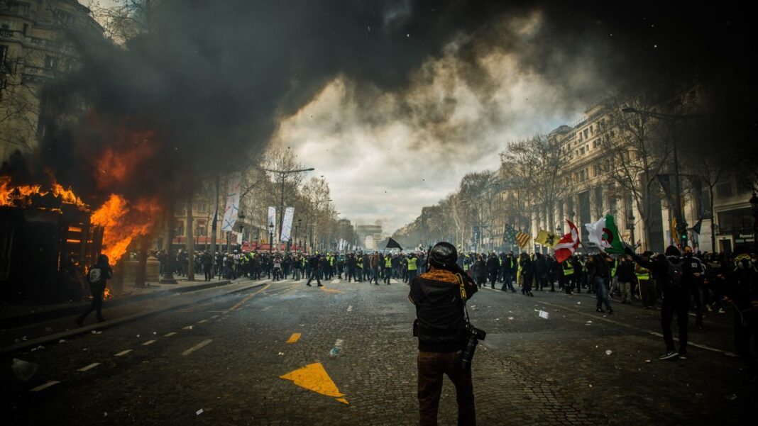 Multitud de manifestantes en una avenida urbana con una gran hoguera y humo negro a la izquierda, un fotógrafo de espaldas en primer plano y edificios a ambos lados.