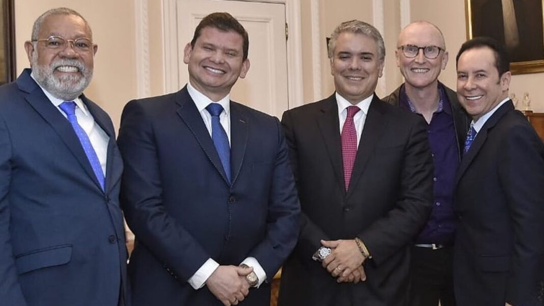 Cinco hombres sonrientes posan juntos en interiores; varios visten trajes oscuros y corbatas, y algunos llevan gafas.